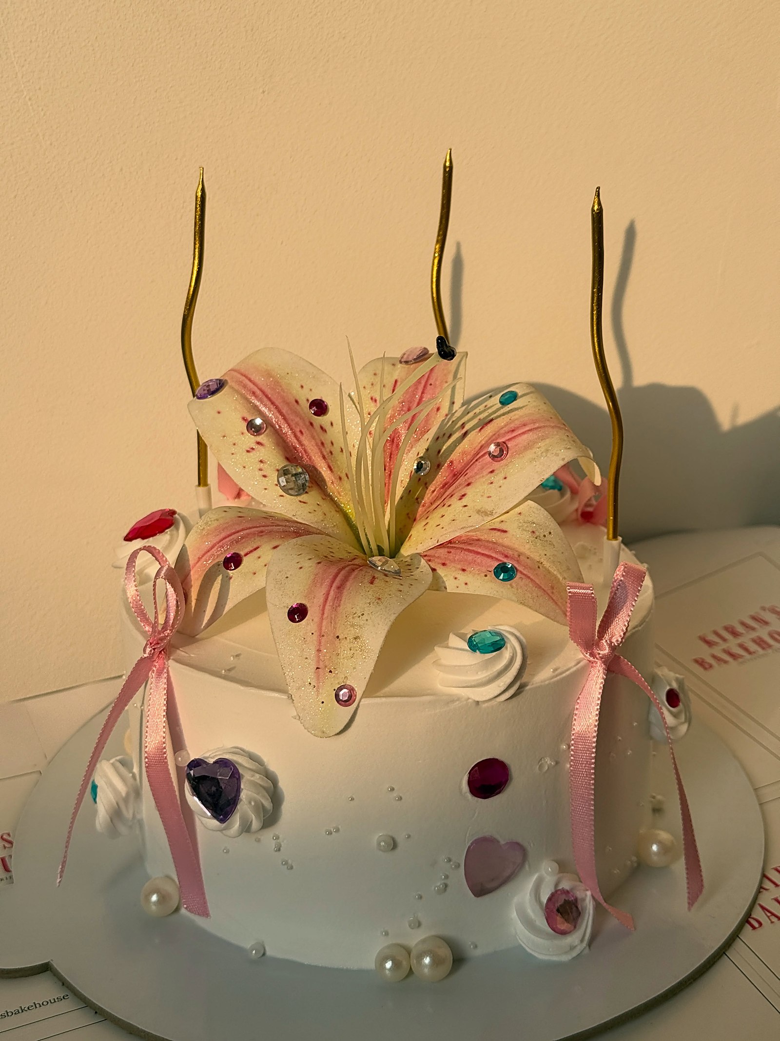 White celebration cake with handmade lily, pink bows and gold candles